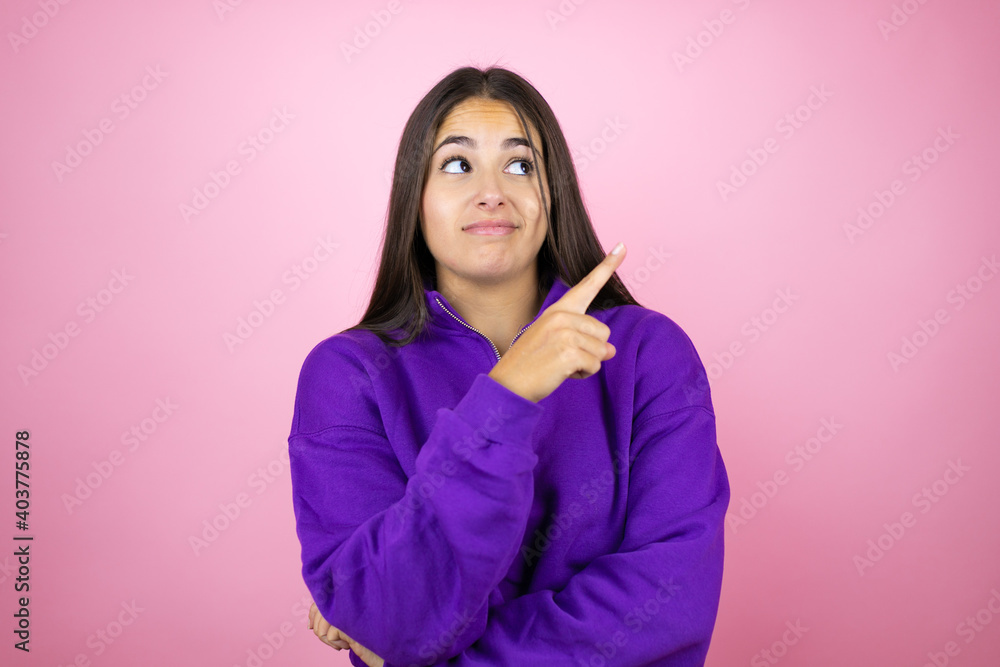 Young beautiful woman wearing sweatshirt over isolated pink background confused and pointing with hand and finger to the side Young beautiful woman wearing sweatshirt over isolated pink background confused and pointing with hand and finger to the side