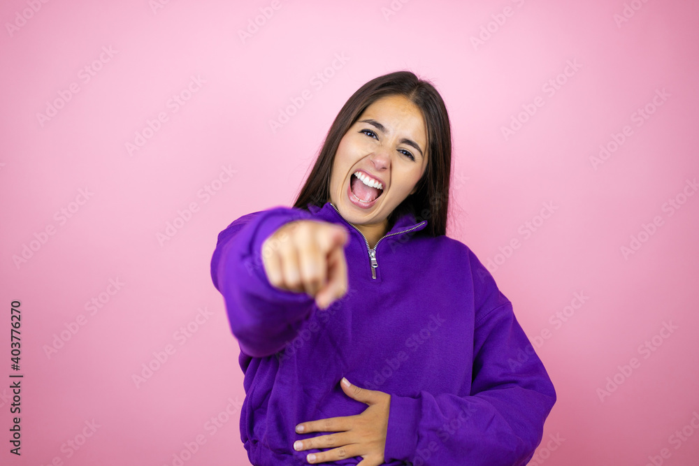 Young beautiful woman wearing sweatshirt over isolated pink background laughing at you, pointing finger to the camera with hand over body, shame expression Young beautiful woman wearing sweatshirt over isolated pink background laughing at you, pointing finger to the camera with hand over body, shame expression