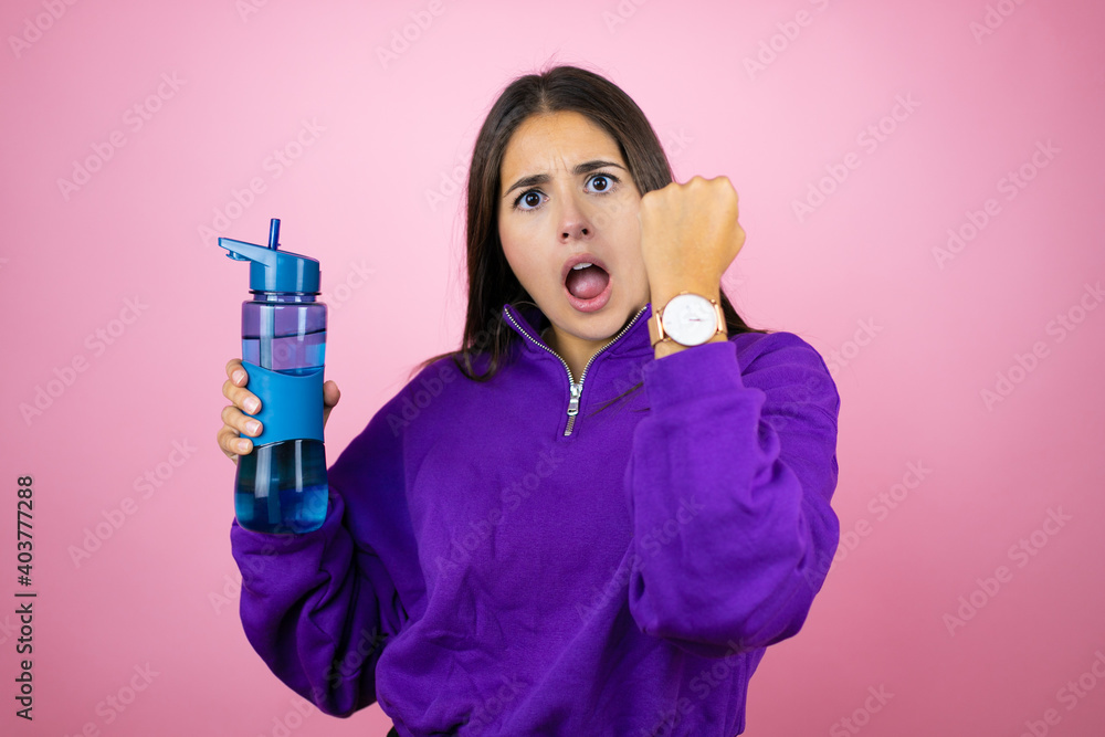 Young beautiful woman wearing sweatshirt holding a water bottle over isolated pink background surprised and showing her watch because it's late Young beautiful woman wearing sweatshirt holding a water bottle over isolated pink background surprised and showing her watch because it's late