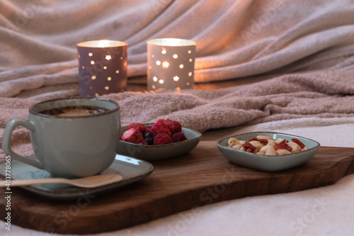 Berries and nuts, cappuccino coffee in a green mug, biodegradable eco spoon on dark wooden cutting board on grey soft cozy hygge plaid blanket. Burning candles