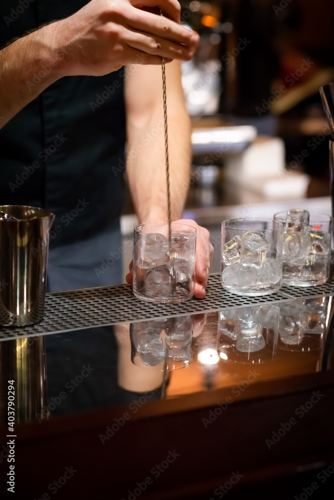 male bartender's hands mixing ice cubes with bar spoon to chill cocktail glasses