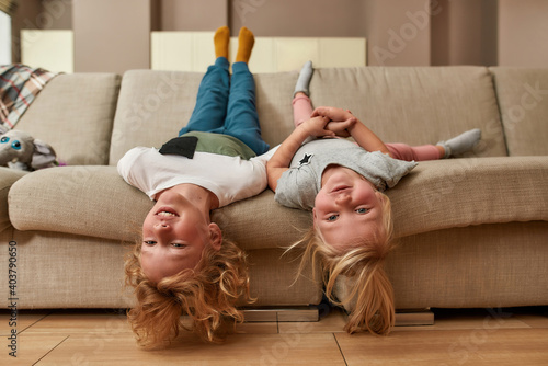 Portrait of playful kids, little boy and girl smiling while lying upside down on a sofa in the living room