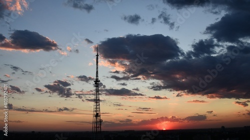 Cloudscape with industrial communication tower and dark purple clouds while golden sunbeams shining through. Red sun cloudscape of sunset. Dark dramatic sky sundown