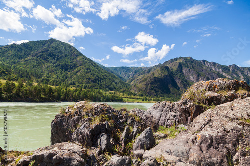 The bank of the Katun river with water containing turquoise clay and a sandy beach, rocky coast against the backdrop of mountains covered with green forest and blue sky with white clouds.