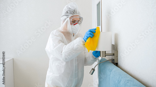 Housekeeper or maid cleaning and desinfecting furniture in hotel room. Person wearing protective medical suit doing cleanup at home photo