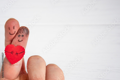 Close-up of two crossed fingers with drawn happy faces holding a small red paper heart on a white wooden background with copy space for text. The concept of love, care, valentine's day.