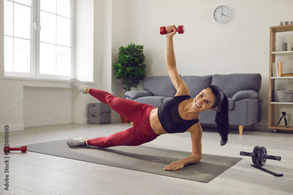 Fototapeta premium Home training. Athletic woman trains lying sideways on a sports mat and lifts up her leg and dumbbell. Athlete does not miss training while at home. Fitness, sport, training and lifestyle concept.
