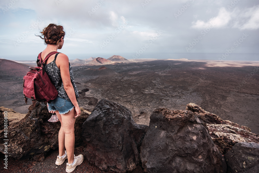 Naklejka premium Young woman with a backpack contemplating the volcanic area Timanfaya Natural Park, in Lanzarote, in Lanzarote