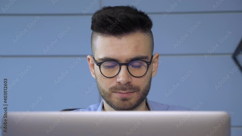 Close Up of A Young European Man with Glasses A Businessman Looking at a Monitor on a Laptop Online in a Bright Cozy Room Home Office Close Up of A Young European Man with Glasses A Businessman Looking at a Monitor on a Laptop Online in a Bright Cozy Room Home Office
