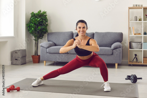 Happy pretty young woman athlete in sportswear doing workout with squats for legs on fitness mat at home with room interior at background. Active healthy lifestyle, training at home during quarantine