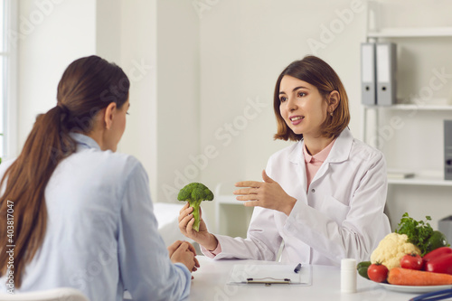 Qualified female dietitian, nutritionist, healthy food expert, doctor of alternative medicine, consultant in health center holding broccoli and telling young woman about benefits of eating vegetables photo