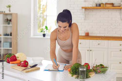 Cheerful pretty slim female athete in sportswear standing in kitchen in sportswear and writing down healthy recipe or daily ration diet at home. Active healthy lifestyle, clean eating concept