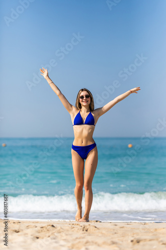Beautiful young woman with raised hands, looking at ocean. Freedom concept, holiday, beach, clear sky background.