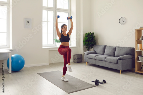 Fototapeta Naklejka Na Ścianę i Meble -  Fit beautiful young female in sportswear having aerobic workout at home. Happy smiling positive woman in sports bra and leggings doing exercises with dumbbells, standing on rubber mat in living-room