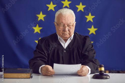 Canvas Print European Court of Justice aka ECJ: Strict serious old judge reading document while sitting at table in courtroom against EU flag background