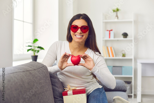 Video conference view of happy smiling young woman in heart-shaped sunglasses holding red heart sitting on sofa with present. Sharing love, getting and giving romantic gifts on Saint Valentine's Day