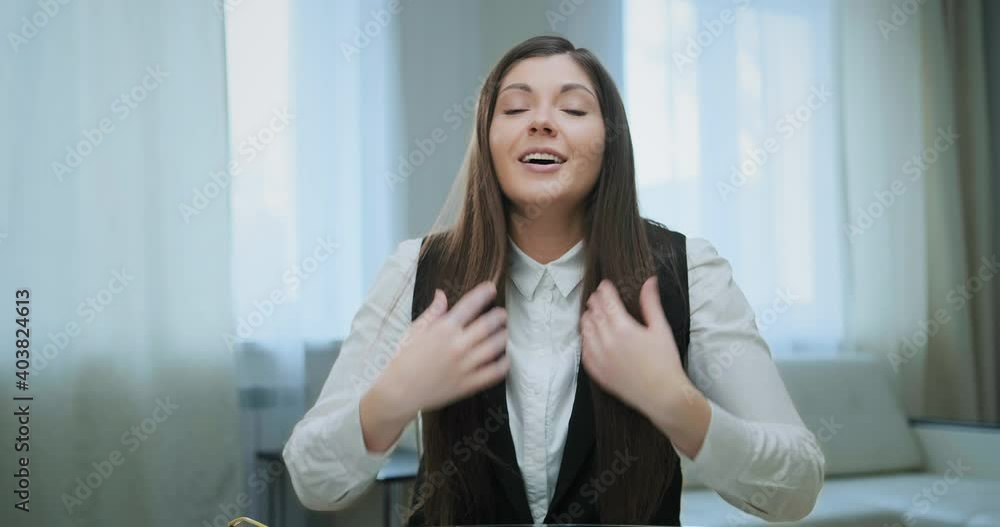 Young brunette fixes long loose hair and talks to corporate manager sitting on white sofa at home at online video conference Young brunette fixes long loose hair and talks to corporate manager sitting on white sofa at home at online video conference