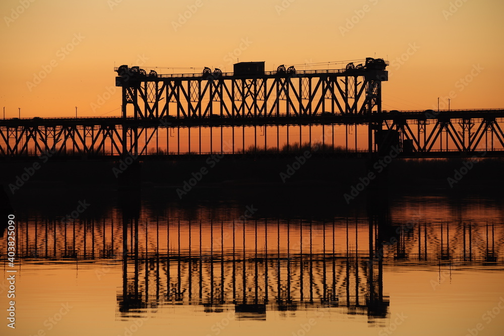 Fototapeta premium Traffic on the bridge over the Dnieper river against the background of an orange sunset. Kryukovsky road-rail bridge in Kremenchug, Ukraine. Industrial silhouette against the background of a natural