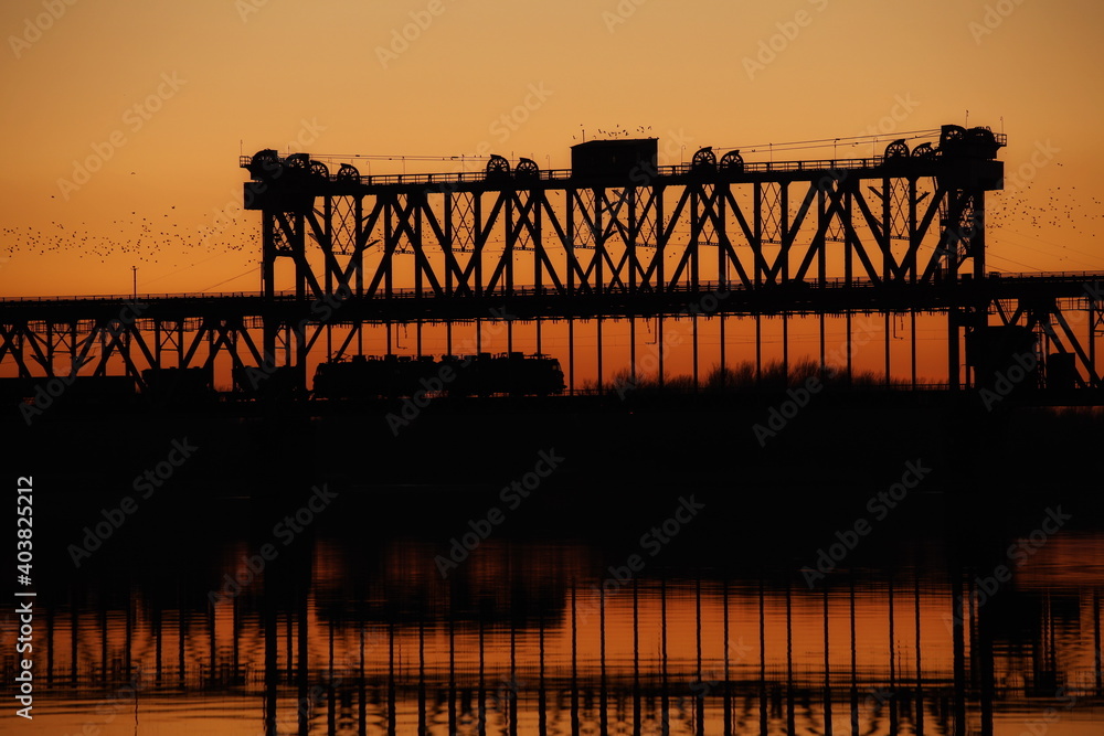 Traffic on the bridge over the Dnieper river against the background of an orange sunset. Kryukovsky road-rail bridge in Kremenchug, Ukraine. Industrial silhouette against the background of a natural
