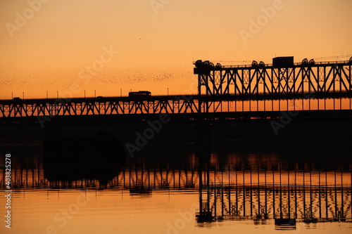 Traffic on the bridge over the Dnieper river against the background of an orange sunset. Kryukovsky road-rail bridge in Kremenchug, Ukraine. Industrial silhouette against the background of a natural