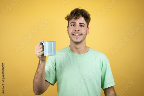 Handsome man wearing a green casual t-shirt over yellow background enjoying and drinking a cup of coffee