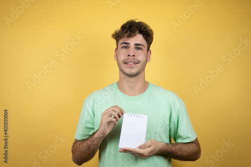 Handsome man wearing a green casual t-shirt over yellow background smiling and showing blank notebook