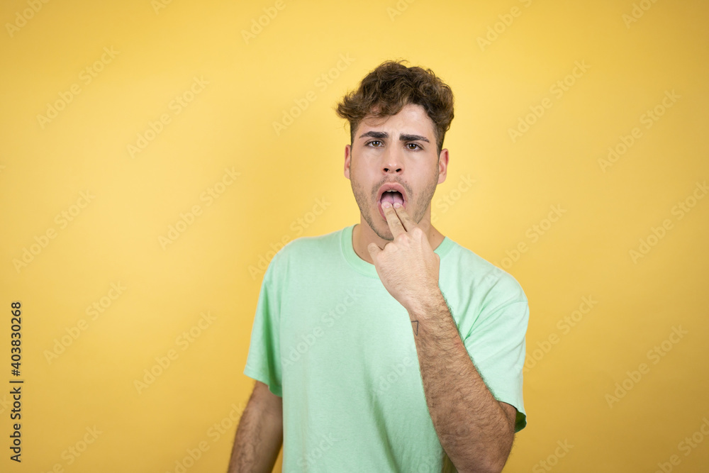 Handsome man wearing a green casual t-shirt over yellow background disgusted with his hand inside his mouth pretending he is going to vomit Handsome man wearing a green casual t-shirt over yellow background disgusted with his hand inside his mouth pretending he is going to vomit