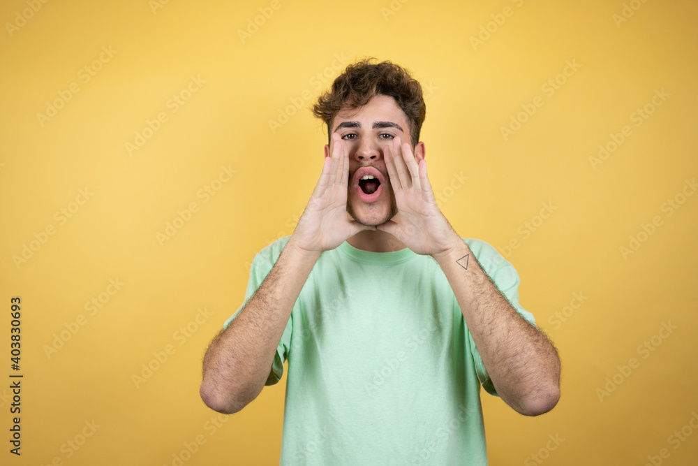 Handsome man wearing a green casual t-shirt over yellow background shouting and screaming loud to side with hands on mouth Handsome man wearing a green casual t-shirt over yellow background shouting and screaming loud to side with hands on mouth