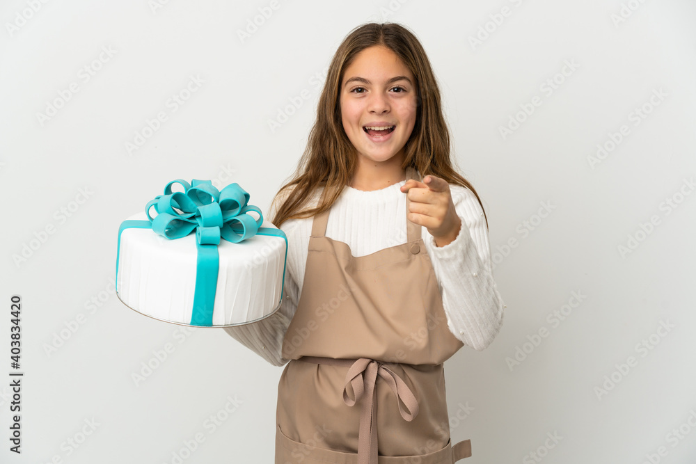 Little girl holding a big cake over isolated white background surprised and pointing front Little girl holding a big cake over isolated white background surprised and pointing front