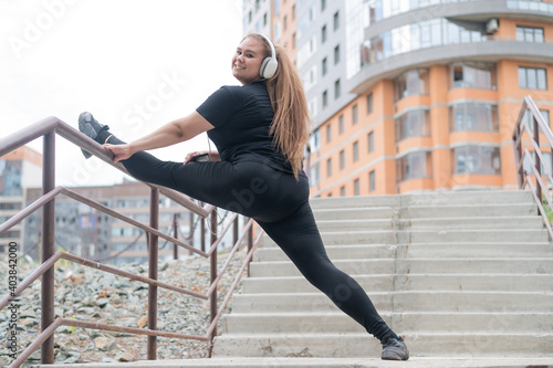 Fat young woman doing stretching exercises and listening to music with headphones outdoors
