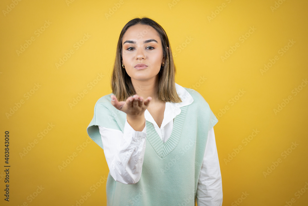 Pretty young woman standing over yellow background looking at the camera blowing a kiss with hand on air being lovely and sexy. Love expression. Pretty young woman standing over yellow background looking at the camera blowing a kiss with hand on air being lovely and sexy. Love expression.