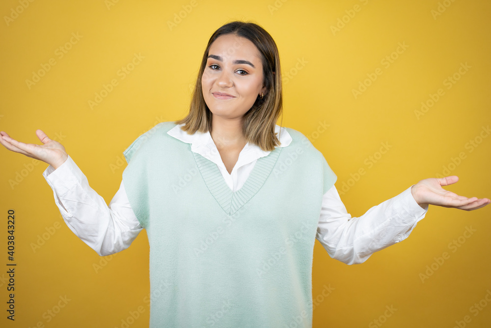 Pretty young woman standing over yellow background clueless and confused expression with arms and hands raised Pretty young woman standing over yellow background clueless and confused expression with arms and hands raised