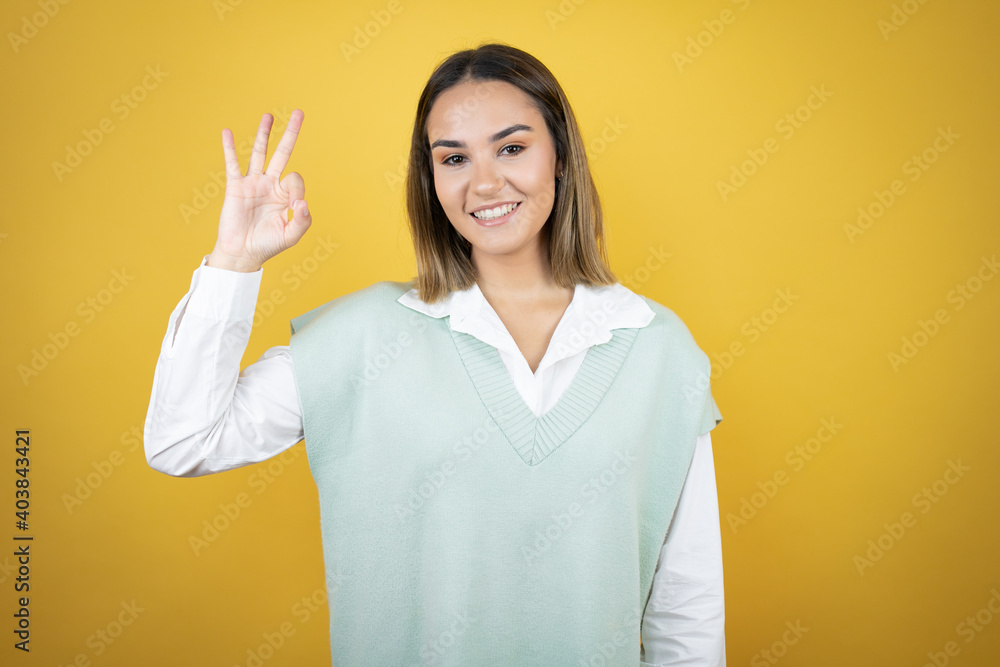 Pretty young woman standing over yellow background doing ok sign with fingers and smiling, excellent symbol Pretty young woman standing over yellow background doing ok sign with fingers and smiling, excellent symbol
