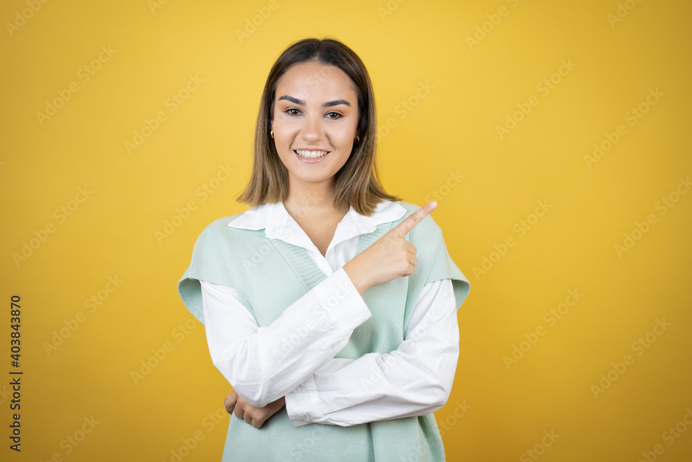Pretty young woman standing over yellow background smiling confident pointing with hand and finger to the side Pretty young woman standing over yellow background smiling confident pointing with hand and finger to the side