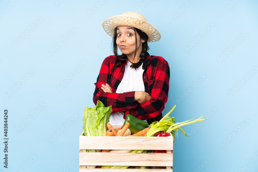 Young farmer Woman holding fresh vegetables in a wooden basket making doubts gesture while lifting the shoulders
