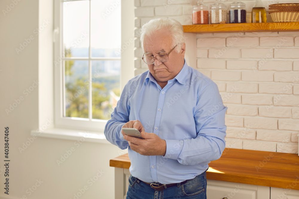 Senior man looks at smartphone and reads the news intently while standing in the kitchen at home. Concept of using digital technologies by mature people in everyday life.