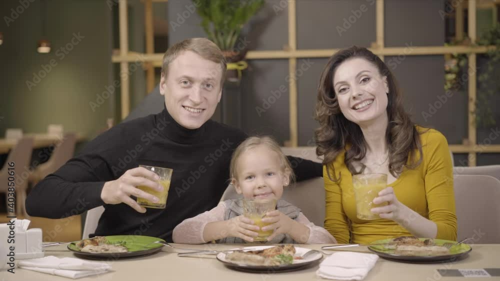 Happy young family clinking glasses with orange juice in pizza restaurant and smiling at camera. Portrait of cheerful Caucasian mother, father, and cute little daughter resting in pizzeria. Happy young family clinking glasses with orange juice in pizza restaurant and smiling at camera. Portrait of cheerful Caucasian mother, father, and cute little daughter resting in pizzeria.