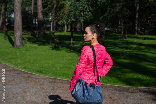 A young woman with a sports bag on her shoulder stands in the park. Side view. © Taras Grebinets