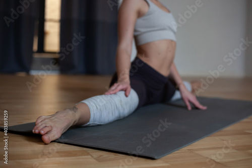 no face young woman doing twine stretching on youga mat in room with wooden floor and white walls. Selective focus
