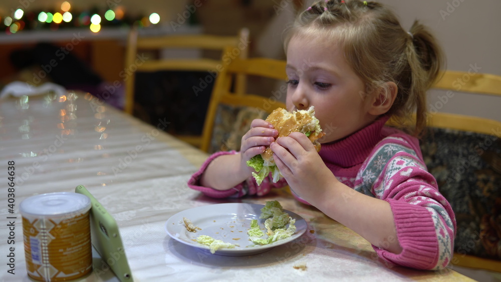 Little girl eating hamburger. Child with hamburger watching cartoons on smartphone Little girl eating hamburger. Child with hamburger watching cartoons on smartphone