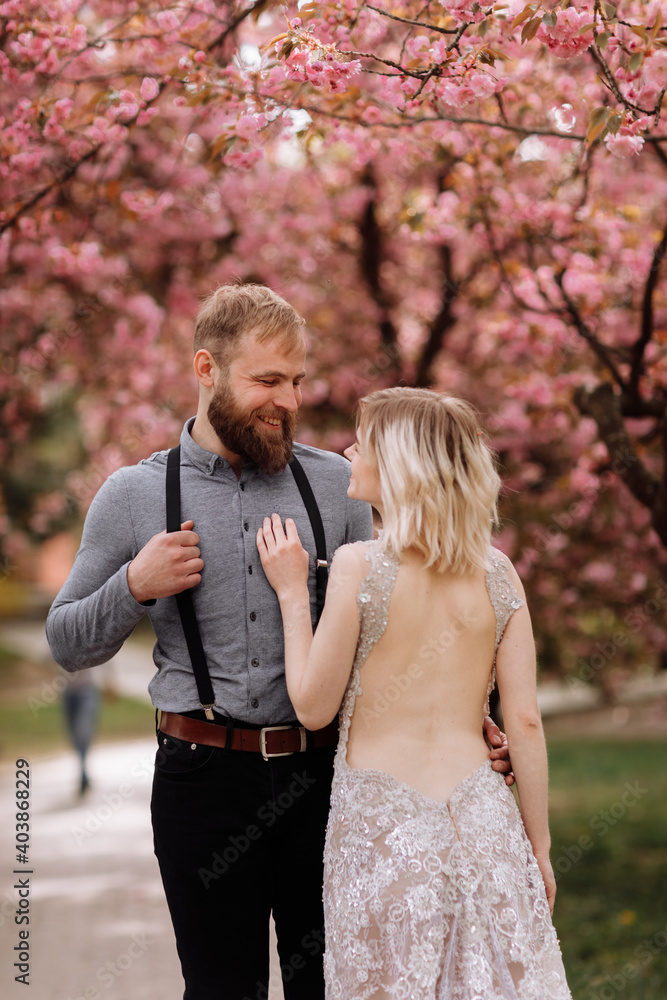 Fototapeta premium Beautiful, cheerful and lively newlyweds, groom and bride are hugging near the blooming pink cherry blossom. Wedding portrait of a close-up of a smiling bearded groom and a cute bride.