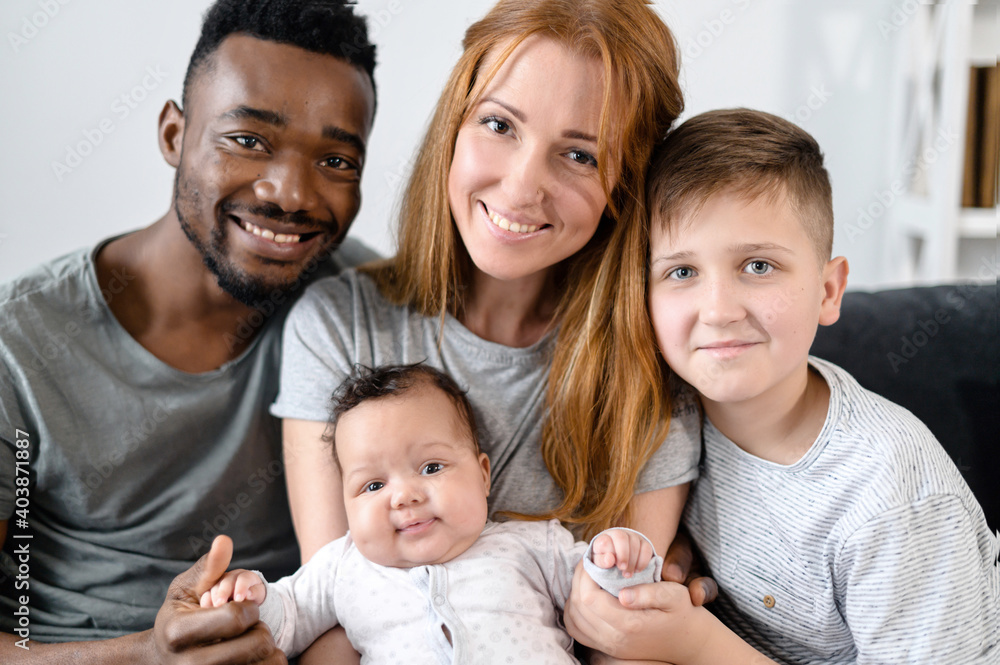 A portrait of happy multiracial family at home. A multiethnic spouses, their cute newborn baby girl and school-age son look into the camera and smile. A portrait of happy multiracial family at home. A multiethnic spouses, their cute newborn baby girl and school-age son look into the camera and smile.