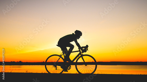 Unrecognizable silhouette man riding bicycle against sunset sky. Road biking cyclist workout, riding racing bicycle on open road. Workout for triathlon. Dramatic sunset background. Copy space
