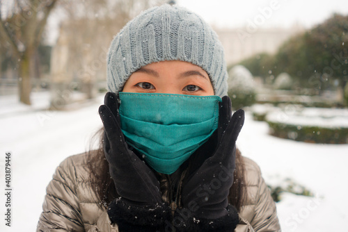 snowfall in covid19 time - young happy and attractive Asian Japanese woman in winter hat and mask at beautiful city park playing with snow cheerful enjoying cold Christmas holidays