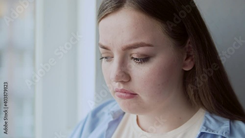 Sad chubby girl lowering her eyes feeling sadness, lonely standing near window