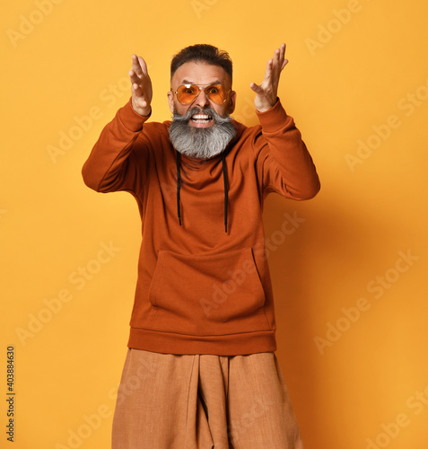 brutal gray-haired bearded hipster in sportswear and fashionable sunglasses is screaming or shouting loudly. Sports fashion, emotions of people. Headshot in closed studio on yellow background
