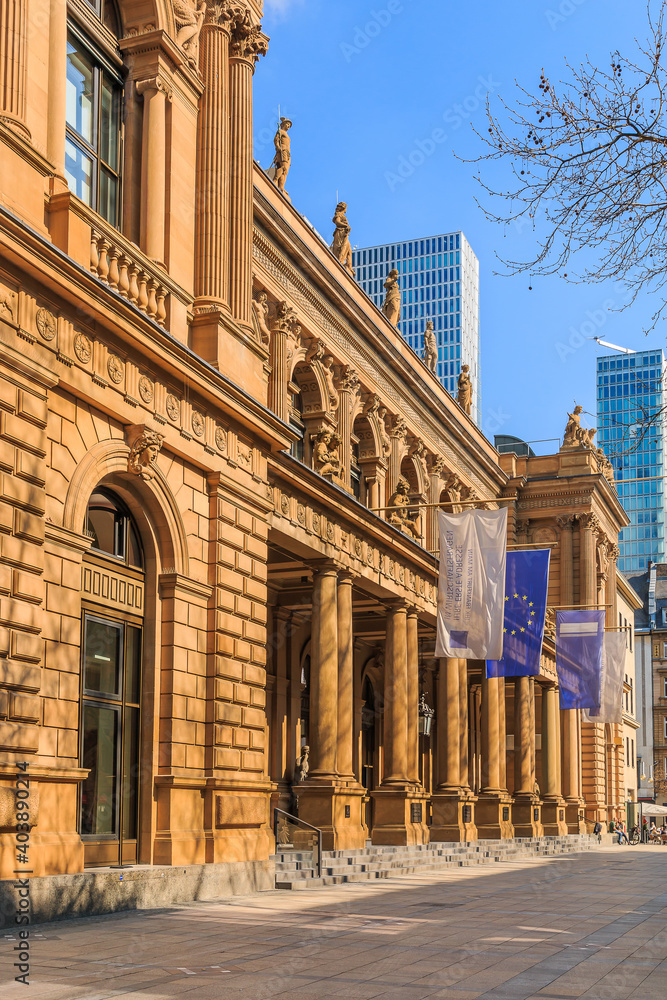 Building of the stock exchange in Frankfurt with flags above entrance. brown colored historical commercial building in baroque style. Skyscrapers in the background. Trees in the foreground in spring