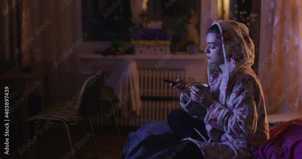 Young woman sitting on bed watching tv and drinking tea Young woman sitting on bed watching tv and drinking tea