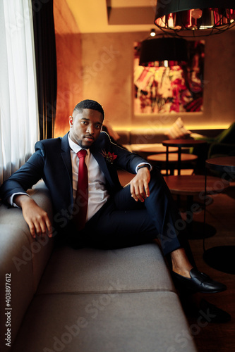 Portrait of handsome African American business male. Attractive charming man look at the camera, smiling, sitting at the sofa. Young adult groom wearing stylish formal suit, excited about marriage