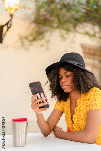 young black girl with curly hair, in yellow dress and black hat, checking her phone sitting in restaurant